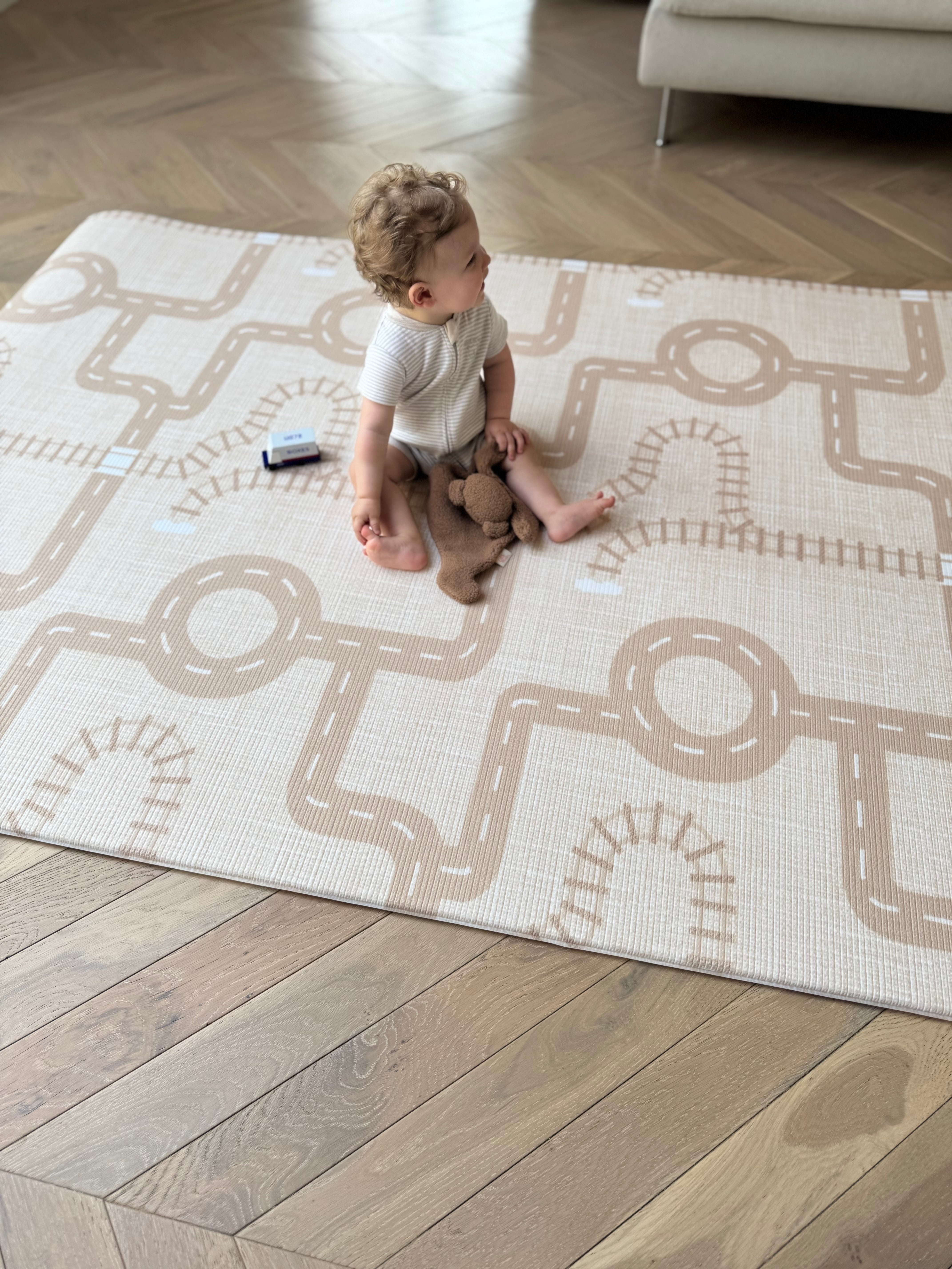  A baby is seated on a rug featuring a train track design, surrounded by colorful toy trains.