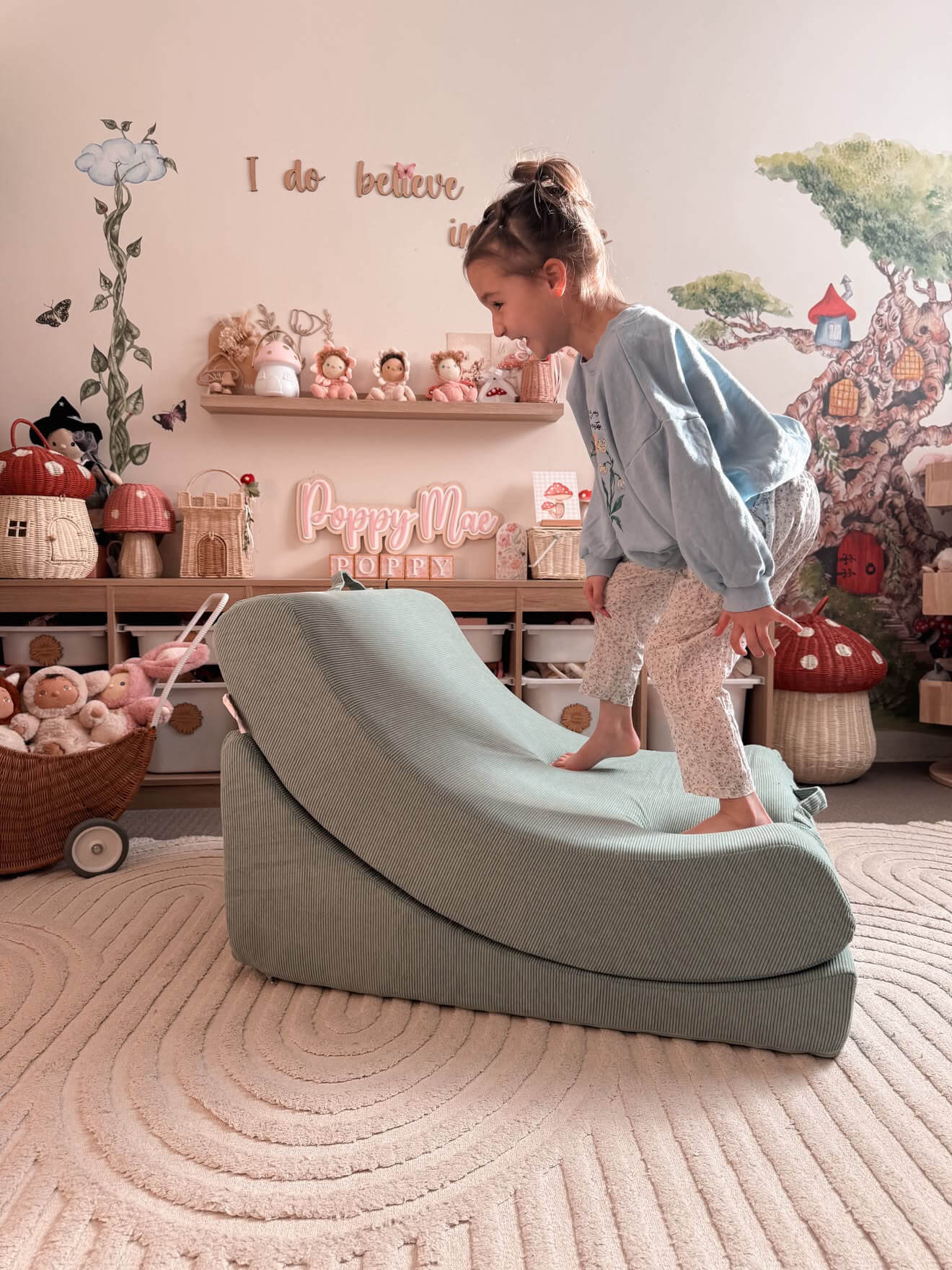 A young girl in a light blue sweatshirt and patterned leggings stands on a curved, wedge-shaped mint Cove Sesnory Lounger memory foam chair in a cozy, decorated children's room.