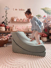 A young girl in a light blue sweatshirt and patterned leggings stands on a curved, wedge-shaped mint Cove Sesnory Lounger memory foam chair in a cozy, decorated children's room.