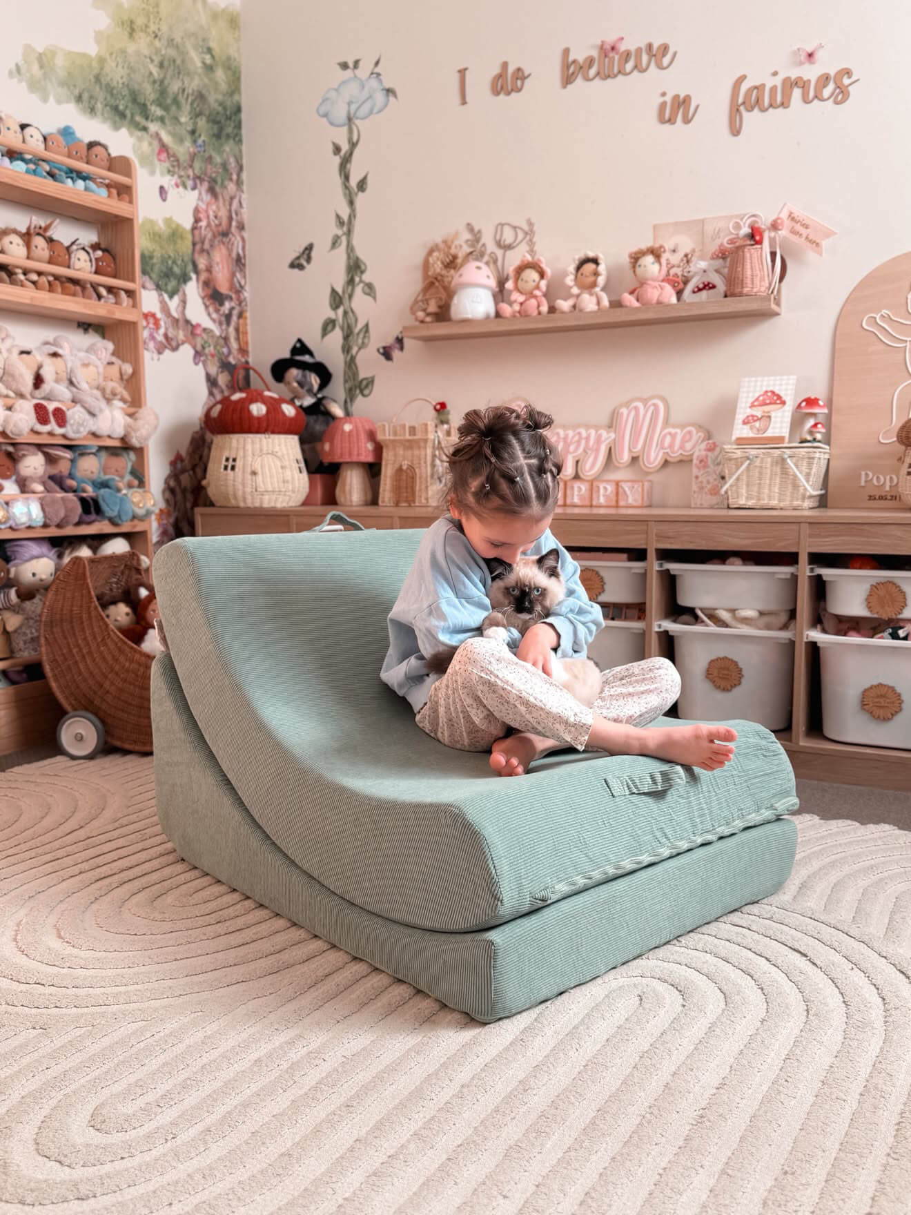 A young child sits on a curved, low-profile lounge chair in a cozy, decorated children's room. The lounge chair is a dusty mint color.
