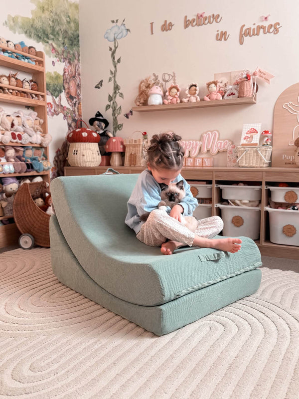 A young child sits on a curved, low-profile lounge chair in a cozy, decorated children's room. The lounge chair is a dusty mint color.