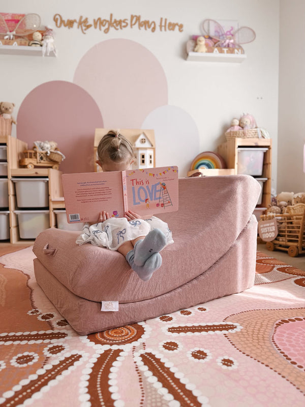 A child sits in a dusty rose crescent-shaped sensory lounge chair, reading a book with their back to the camera in a styled nursery setting.