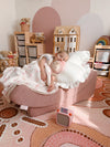 A child lying on a dusty rose crescent-shaped lounger in a decorated nursery, holding a plush toy. The room features a pink and beige patterned rug, wooden toy storage shelves, and a dollhouse.