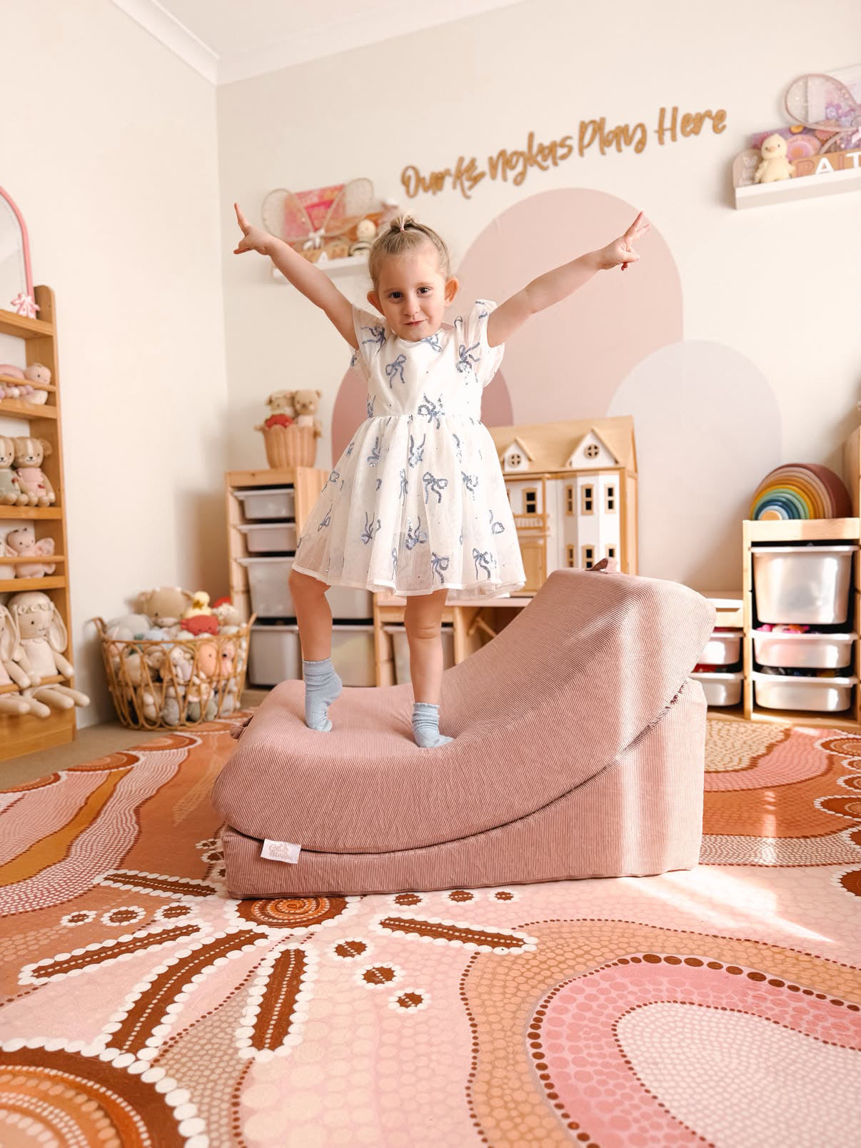 A young child stands on a dusty rose crescent-shaped memory foam sensory chair in a decorated nursery room with a patterned rug, wooden shelves, and toys.