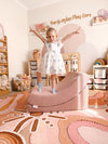 A young child stands on a dusty rose crescent-shaped memory foam sensory chair in a decorated nursery room with a patterned rug, wooden shelves, and toys.
