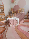 A pink fabric memory foam chair with a white throw blanket draped over it, set in a child's playroom on a mothers love rug with wooden toy shelves and dollhouse in the background.