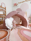 A pink fabric cove sensory lounger positioned as a cubby with a large white ruffled pillow and a small plush doll, set in a decorated children's room with patterned flooring and shelves.