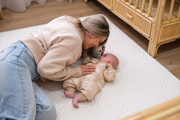 A woman lies on the floor, smiling at a baby who is playing beside her.