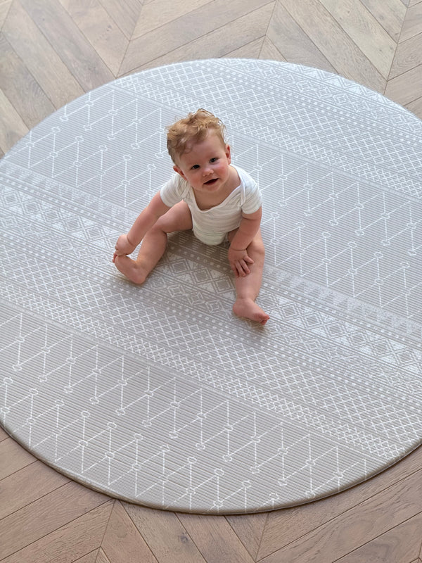 A baby sits happily on a rug with a grey boho pattern