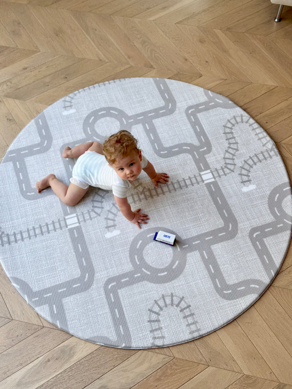 A baby is comfortably resting on a round rug adorned with a car pattern, exploring the soft surface with curiosity.
