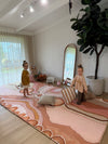 Two children playing on an extra large "Mother's Love" Aboriginal art memory foam mat in a modern room with large windows and decorative plants.