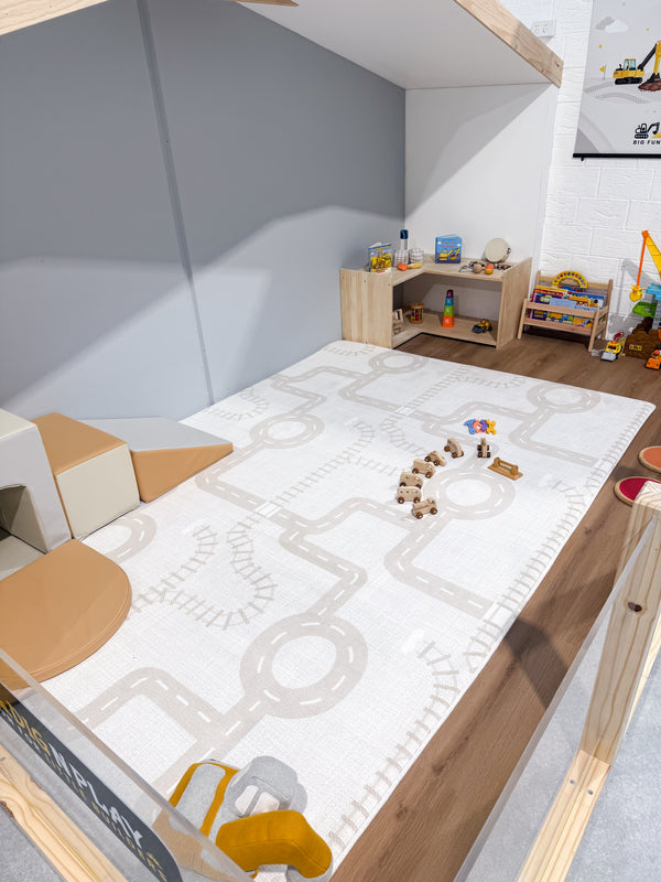 A children's memory foam play mat with a road and track pattern in beige and white, laid out on a wooden play floor in a playroom setting.
