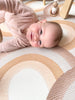 Baby smiling while laying on Archie Retro Large playmat. Side A shows a brown arch pattern.
