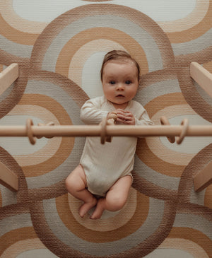 Baby laying on Archie Retro playmat while playing with wooden gym. The playmat has a brown rainbow pattern.